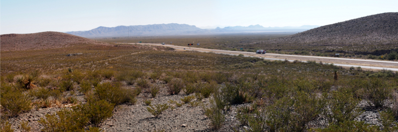 [More sagebrush as the land goes down a hill and off to distant mountains. Across the round is another sagebrush-covered hill. Haze fills the sky. Only a few vehicles on the road.]